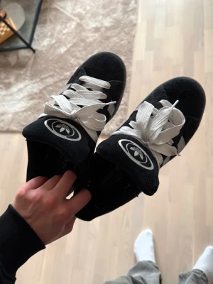 Adidas Campus svarta sneakers - Svarta Adidas Campus sneakers med vita och gråa detaljer och klassiska tre ränder på sidan. Skorna har vit logga på plösen och hälen, samt vita skosnören. Ovandelen är i mocka och sulan är grå. Perfekta för en clean och stilren look.