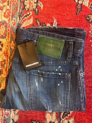 Blå jeans från Dsquared2 - Säljer ett par blå herrjeans med slitningar och färgstänk i storlek 50 från Dsquared2. Sparsamt använda, 9/10 condition. Modellen på bilden är 187.
