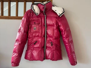 Rosa pufferjacka  - Snygg rosa pufferjacka med glansig finish, huva och svarta detaljer. Jackan har flera fickor med dragkedja och knapp, samt Moncler-logga på bröstet. Perfekt för kalla vinterdagar och riktigt trendig look.