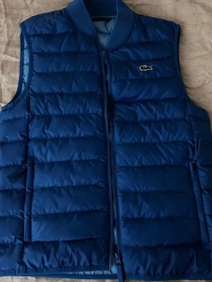 Blå dunväst från Lacoste - Snygg blå dunväst från Lacoste med quiltad design och dragkedja framtill. Västen har ribbad krage och två sidofickor. Ikoniska Lacoste-loggan sitter på bröstet. Perfekt för lager-på-lager och ger en sportig vibe.