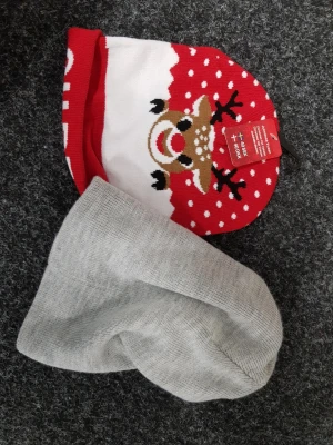 Två mössor, julmotiv & grå basic - Säljer två mössor: en röd och vit med gulligt renmotiv och snöflingor, perfekt för julen, samt en enkel ljusgrå stickad mössa i mjukt material. Båda är bekväma och passar till kalla dagar. Den röda är helt ny medans den andra är lite använd kostar 50kr styck.