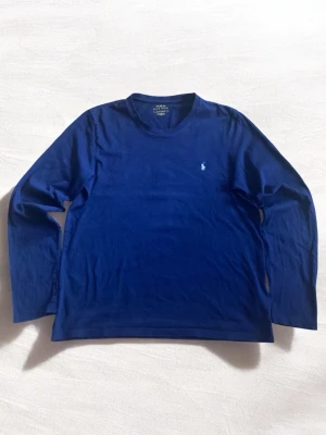 Blå långärmad t-shirt från Polo Ralph Lauren - Snygg marinblå långärmad t-shirt från Polo Ralph Lauren med klassisk rund hals och broderad logga på bröstet. Tillverkad i mjuk bomull som känns skön mot huden. Perfekt för en stilren och avslappnad look.