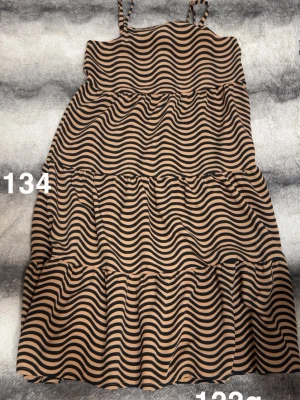  randig klänning - Snygg klänning med vågiga svarta ränder på beige botten. Klänningen har smala axelband och volangdetaljer i flera lager, vilket ger en cool och trendig look. Perfekt för dig som gillar mönster och vill sticka ut.🌸 Använd tre gånger. Storlek 134