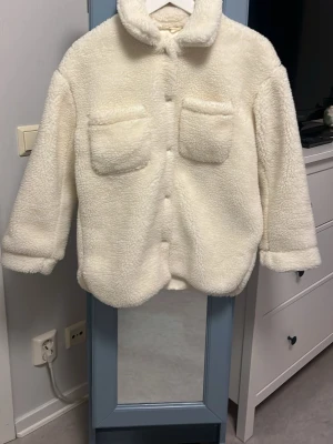 Vit teddyjacka med fickor - Helt oanvänd mysig vit teddyjacka med två stora bröstfickor och knappar framtill. Jackan har en hög krage som kan vikas ner och är gjord i fluffigt, mjukt material som ger en avslappnad och trendig look. Perfekt för kyliga dagar när du vill vara både varm och snygg. BARNSTORLEK 146/152!