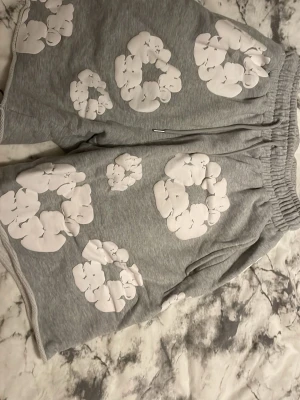 Grå shorts med vita blommor - Grå shorts med elastisk midja och vita blommönster över hela plagget. Mjuk och bekväm passform, perfekta för varma dagar. Sidosömmar och avslappnad stil gör dem lätta att bära till vardags.