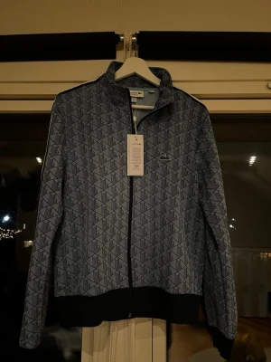 Blå mönstrad ziptröja från Lacoste - Snygg blå ziptröja från Lacoste med grafiskt mönster i svart. Tröjan har ståkrage, dragkedja framtill och svart ribbad mudd vid ärmslut och nederkant. Ikoniska Lacoste-loggan på bröstet. Perfekt för en sportig och trendig look.