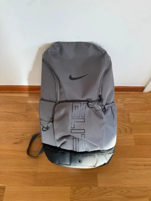 Grå Nike Elite ryggsäck - Snygg grå Nike Elite ryggsäck med svart logga och detaljer. Ryggsäcken har flera fack med dragkedja, vadderade och justerbara axelband samt slitstark botten i svart material. Perfekt för skola, träning eller vardagsbruk.