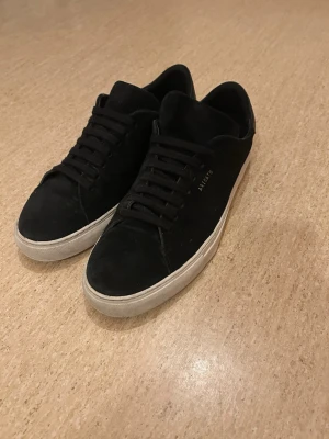 Svarta sneakers från Axel Arigato - Stilrena svarta sneakers från Axel Arigato med ovandel i mocka och klassisk låg siluett. Skorna har svarta skosnören och vit sula som ger en clean kontrast. Diskret logga i guld på sidan. Perfekta för en trendig och avslappnad look.