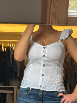 Vit topp - Söt vit linnetopp med volangprydda axelband och knappar framtill. Toppen har en v-ringad urringning och är figurnära i modellen. Perfekt för varma dagar och enkel att styla med jeans eller kjol.