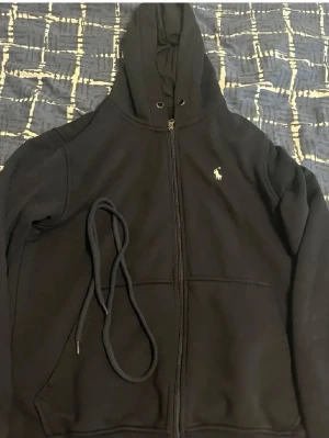 Polo Ralph lauren mörkblå kofta  - Svart hoodie med dragkedja från Polo Ralph Lauren. Klassisk design med huva, dragsko och fickor framtill. Liten vit logga broderad på bröstet. Perfekt för en avslappnad och stilren look.