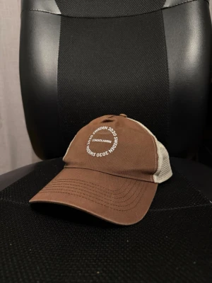 Brun och beige truckerkeps Sweden 2020 - Säljer en brun och beige truckerkeps från ”Consolamini” med vit mesh på sidorna och bak. Framsidan har ett tryck med texten 'Sweden 2020' i vitt. Kepsen har böjd skärm och justerbar snapback. Perfekt för en avslappnad streetwear-look.