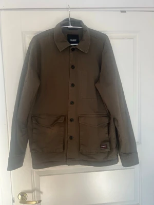 Olivgrön overshirt från Pull&Bear - Stilren olivgrön overshirt från Pull&Bear med klassisk krage och stora framfickor. Skjortjackan har knäppning framtill och en liten logotyp på ena fickan. Perfekt lager-på-lager-plagg för streetwear-stilen.