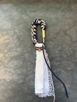Handgjord väsksmycke med spets och rep - Unikt handgjort väsksmycke med flätat rep i marinblått och beige, dekorerat med spets, tygband och en liten guldfärgad stjärnberlock. Perfekt att hänga på väskan för en personlig touch.