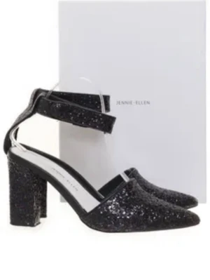 Svarta glittriga pumps Jennie Ellen - Säljer ett par svarta pumps från Jennie Ellen med glittrig yta och bred klack. Skorna har spetsig tå och en bred rem runt ankeln som ger en cool och trendig look. Perfekta för att sticka ut och addera lite extra glam till outfiten. Box förekommer inte!