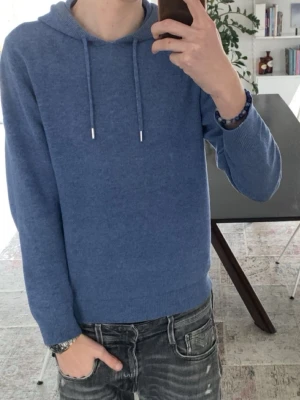 Blå hoodie i kashmir - En stilren blå hoodie i mjukt kashmirtyg med ribbade muddar och huva med dragsko. Perfekt för dig som gillar en enkel men lyxig look. Passar till allt och är superbekväm att bära. Inga synliga loggor eller tryck.