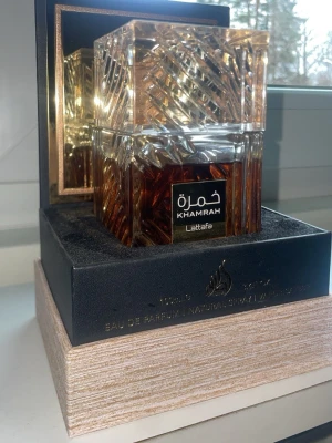 Lattafa Khamrah Eau de Parfum 100ml - Lyxig parfym från Lattafa, Khamrah, i en elegant fyrkantig glasflaska med vackert mönstrad design. Flaskan har en varm bärnstensfärgad vätska och levereras i en exklusiv presentförpackning. Volymen är 100 ml och det är en Eau de Parfum.