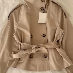 Kort beige trenchcoat från Bershka - Stilren kort trenchcoat i beige från Bershka med dubbelknäppning, axelklaffar och knytskärp i midjan. Jackan har bred krage, dekorativa knappar och justerbara ärmslut. Perfekt för dig som gillar klassisk men trendig look. Helt ny i sin förpackning. Storlek S