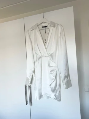 Vit omlottklänning från ZARA - Säljer en stilren vit omlottklänning från ZARA med lång ärm och v-ringad hals. Klänningen har snygga draperingar i sidan och breda manschetter. Perfekt för dig som gillar minimalistisk och trendig stil. Helt oanvänd, har prislapp kvar. 