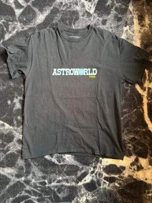 Astroworld Tour svart t-shirt - Säljer en svart Astroworld Tour t-shirt med tryck fram och bak. Framsidan har Astroworld-loggan i blått och gult, baksidan har ett stort motiv av Travis Scott och turnédatum. Klassisk passform och rund hals, perfekt för dig som gillar streetwear.