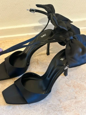 Svarta pumps med rosett från Zara - Säljer ett par svarta pumps från Zara med hög klack, öppen tå och bred rem över foten. Skorna har en elegant rosett vid ankeln och smal ankelrem med spänne. Använd 1 gång.