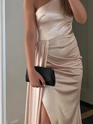 Champagnefärgad balklänning - Snygg champagnefärgad klänning i satin med en axel, hög slits och draperad midja. Klänningen har en elegant cut-out detalj vid midjan och en figurnära passform som ger en lyxig känsla. Lite lång på mig som är 170. 