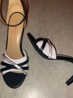 Svarta klackskor med glitterdetalj - Säljer ett par snygga och bekväma klackskor från Catwalk med svarta och glittriga remmar över foten. Skorna har öppen tå, smal klack på ca 6-8cm och justerbart spänne runt ankeln. Det vita på bilden är alltså bara ”skyddet” som följer med skorna. Sparsamt använd, inga synliga defekter så vitt jag vet. Kan självklart diskutera pris🩷