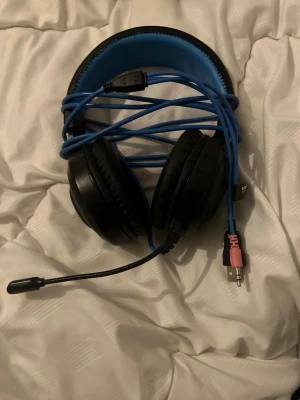 Svarta gaminghörlurar med blå detaljer - Säljer ett par over-ear gaminghörlurar med svart och blå design. Hörlurarna har mjuka, vadderade kåpor och ett justerbart huvudband i konstläder. Utrustade med mikrofon och flera anslutningar: USB, 3.5mm och 2x 3.5mm-kontakter. Perfekta för gaming och voicechatt.