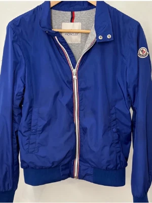 Blå vindjacka från Moncler - Snygg blå vindjacka från Moncler med vit och röd dragkedja, ribbade muddar och Moncler-logga på ärmen. Jackan har ståkrage med tryckknappar och två fickor framtill. Tillverkad i lätt och slitstarkt material, perfekt för blåsiga dagar.