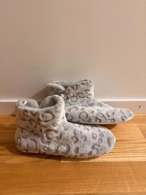 Grå fluffiga tofflor med leopardmönster - Mysiga grå tofflor i fluffigt material med leopardmönster och små bollar som dekoration. De har en högre ankelmodell och är perfekta för att hålla fötterna varma hemma. Supermjuka och sköna att ha på sig. Från Primark, aldrig använda 