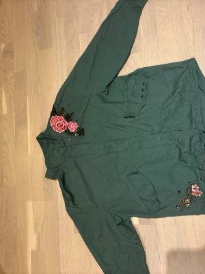 Grön parkas med blommiga broderier - Stilren grön parkas med snygga broderade rosor i rosa och rött på axeln och nedtill. Jackan har dragsko i midjan, två fickor framtill och knappar vid ärmslut. Materialet är bomullsliknande och jackan har en avslappnad passform. Perfekt för dig som vill ha en unik touch på ytterplagget.