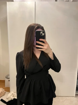 Svart omlottblus med markerad midja - Snygg svart omlottblus från Shein med långärmad design och markerad midja. Blusen har v-ringning och ett lätt utställt peplumformat avslut nedtill. Perfekt att styla med både jeans och kjol för en trendig look.