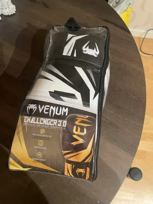 Venum Challenger 3.0 boxningshandskar - Säljer ett par Venum Challenger 3.0 boxningshandskar i svart och vitt med coola grafiska detaljer. Handskarna är tillverkade i syntetiskt läder och har en robust konstruktion med bred kardborre för extra stöd. Perfekt för träning och sparring.
