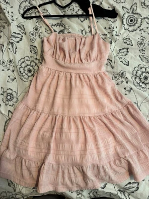 Ljusrosa volangklänning  - Supersöt ljusrosa klänning med tunna axelband och smock i ryggen. Klänningen har volanger och ett strukturerat tyg som ger en romantisk vibe. Perfekt för varma dagar och har en luftig, flowy passform. Materialet känns som bomull eller en bomullsblandning.