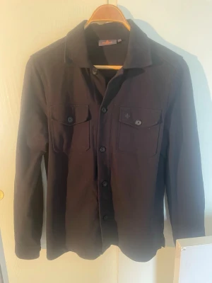  overshirt från Morris - Stilren svart overshirt från Morris med klassisk krage och två bröstfickor med knappstängning. Skjortjackan har raka ärmar och knappar framtill. Perfekt att bära som lager över t-shirt eller tunn tröja. Materialet känns mjukt och har en matt finish.     Skriv gärna om ni vill se hur den sitte eller om jag har övriga frågor
