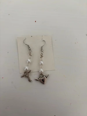 Örhängen med pärlor och berlocker - Säljer ett par unika örhängen med vita pärlor och silverfärgade berlocker – ena med en nyckel och den andra med ett hjärta med vingar och lås. Perfekta för dig som gillar detaljer och vill sticka ut lite extra.