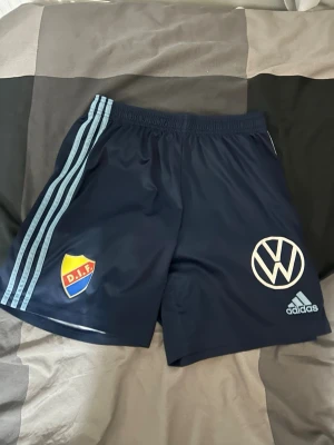 Blå Djurgården shorts från Adidas - Mörkblå sportshorts från Adidas med ljusblå ränder längs sidorna. Djurgårdens IF-märke på ena benet och Volkswagen-logga samt Adidas-logga på det andra. Perfekta för träning eller match.