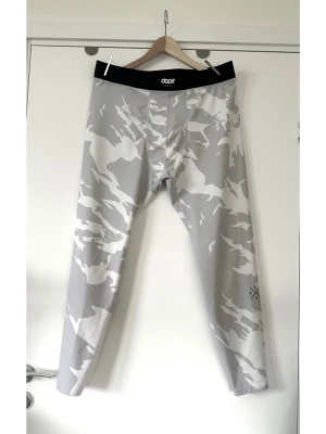 Gråvita camo leggings från Dope, storlek L - Grå-vita träningsbyxor / tights /underställ från DOPE i storlek L. Populära ”Snuggle” - modellen med coolt kamouflagemönster och svart linning med Dope-logotypen framtill och en bergslogotyp baktill. En ”DOPE”-logotyp i form av ett kors finns på höger ben. 30% spandex, så bra medföljande material som passar för även intensiv träning. Nypris 700:-. Använd 1 gång, så i mycket gott nyskick. 