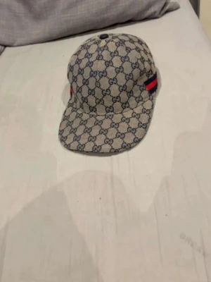 Gucci keps med GG-mönster - Säljer en snygg keps från Gucci med klassiskt GG-monogram i blått på beige botten. Kepsen har böjd skärm och en röd-blå rand på sidan som ger extra lyxig känsla. Perfekt accessoar för dig som gillar streetwear och designerplagg.