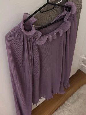 Plisserad lila blus med volangkrage - Säljer en lila plisserad blus med långa ärmar och volangdetaljer vid halsringningen. Blusen har en lös passform och är tillverkad i ett lätt, flowy material som ger ett snyggt fall. Perfekt för dig som gillar trendiga detaljer och en unik look.