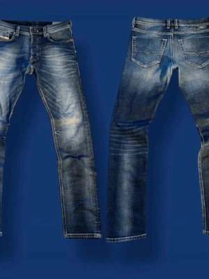 Blå slitna jeans från Diesel - Säljer ett par blå jeans från Diesel med snyggt sliten tvätt och detaljerade sömmar. klassiska fem fickor och Diesel Industry-märke på myntfickan. Perfekta för en avslappnad och trendig look.
