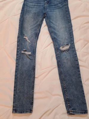Pull&Bear Blå slitna skinny jeans - Säljer ett par blå skinny jeans från Pull&Bear med slitningar och hål på båda benen. Jeansen har klassisk femficksdesign och är tillverkade i stretchigt denimtyg för en tajt passform. Perfekta för en avslappnad och trendig look. I stort sett som nya.