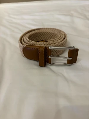 Beige flätat bälte med skinn-detaljer - Snyggt beige flätat bälte med bruna skinn-detaljer och silverfärgat spänne. Perfekt accessoar för att lyfta din outfit med en stilren touch. Passar till både jeans och chinos.