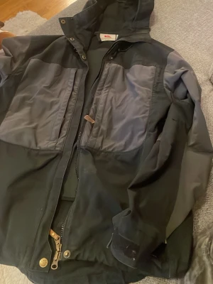 Svart och grå vindjacka Fjällräven - Snygg svart och grå vindjacka från Fjällräven med huva och dragkedja. Jackan har två stora bröstfickor med dragkedja och detaljer i läder. Tillverkad i slitstarkt material, perfekt för utomhusaktiviteter. Klassisk design med tryckknappar och justerbar nederkant.