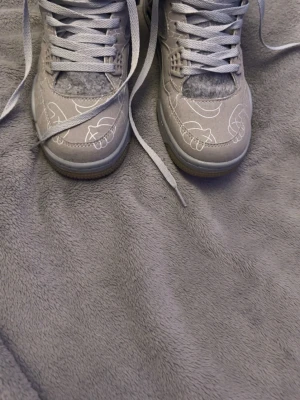 Gråa Air Jordan 4 x KAWS sneakers - Unika gråa Air Jordan 4 x KAWS sneakers med mocka och filtliknande detaljer. Skorna har vita KAWS-grafiska mönster på ovandelen, klassisk snörning och synlig Air-enhet i sulan. Perfekta för dig som gillar streetwear och exklusiva samarbeten.