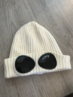Vit ribbstickad mössa från C.P. Company - Säljer en vit ribbstickad mössa från C.P. Company med ikoniska svarta glasögonlinser framtill. Mössan är tillverkad i mjuk ull och har en uppvikt kant. 