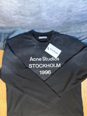 Svart långärmad topp Acne Studios - Cool svart långärmad topp från Acne Studios med tryck i vitt framtill: 'Acne Studios STOCKHOLM 1996'. Rund halsringning och relaxed fit. Perfekt för dig som gillar minimalistisk och stilren design.