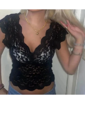 Svart spetsblus med v-ringning - Snygg svart blus i spets med djup v-ringning och korta ärmar. Blusen är genombruten och har vågiga kanter nertill och vid ärmslut. Perfekt för dig som vill ha en feminin och trendig topp.