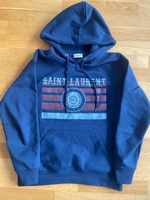 Blå Saint Laurent hoodie med tryck - Snygg blå hoodie från Saint Laurent med huva och dragsko. Framsidan har ett vintageinspirerat tryck med texten 'SAINT LAURENT' och röda samt vita ränder. Stor magficka framtill och ribbade muddar. Perfekt för en avslappnad och trendig look.