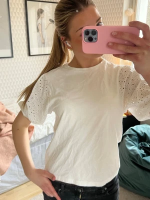 Vit topp med broderade puffärmar - Säljer en söt vit topp med puffiga, broderade kortärmar i bomull. Ärmarna har fina spetsdetaljer som ger en romantisk vibe. Perfekt till jeans eller kjol för en fräsch look. Rund halsringning och normal passform.