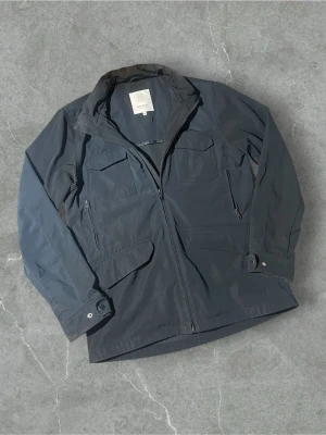 Mörkblå vindjacka från Elvine | L - Stilren mörkblå vindjacka från Elvine med dragkedja framtill, fyra fickor med lock och knappdetaljer vid ärmsluten. Jackan har hög krage och är tillverkad i ett lätt, vindtätt material som passar perfekt för blåsiga dagar.